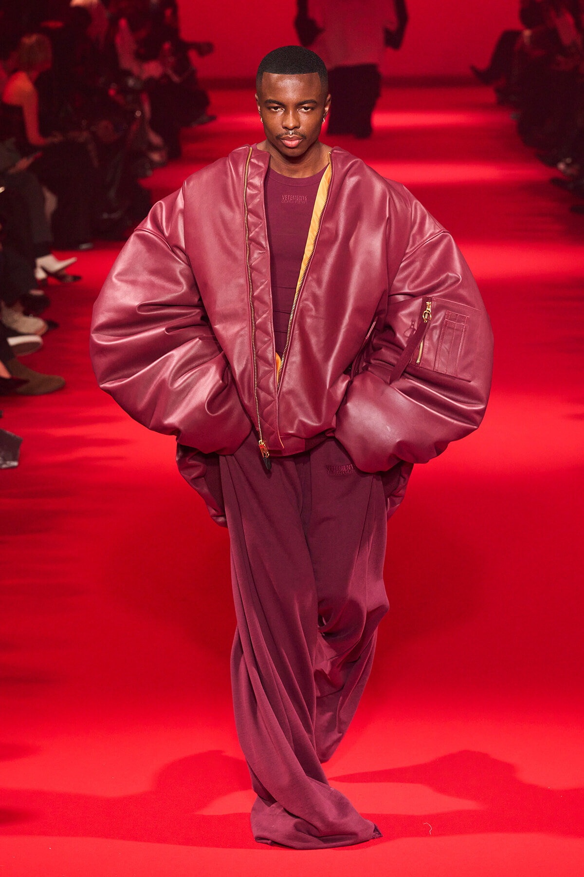Male model on a red-lit runway wearing an oversized burgundy puffer jacket and matching pants, hands in pockets in a fashion show setting.