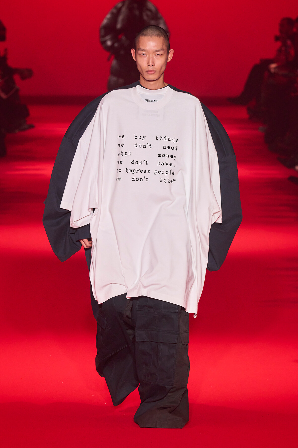 Runway model wearing an oversized white T‑shirt with typed text and black cargo pants on a red catwalk backdrop.