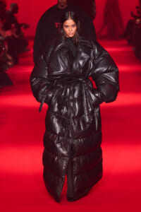 Model walks a red-lit runway wearing a oversized black puffer coat with a belt tied at the waist and large shoulders.