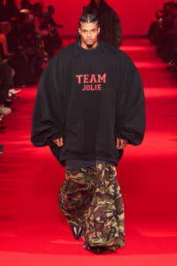 Male model walking a red runway, wearing an oversized black sweatshirt with the words 'TEAM JOLIE' and camo cargo pants.