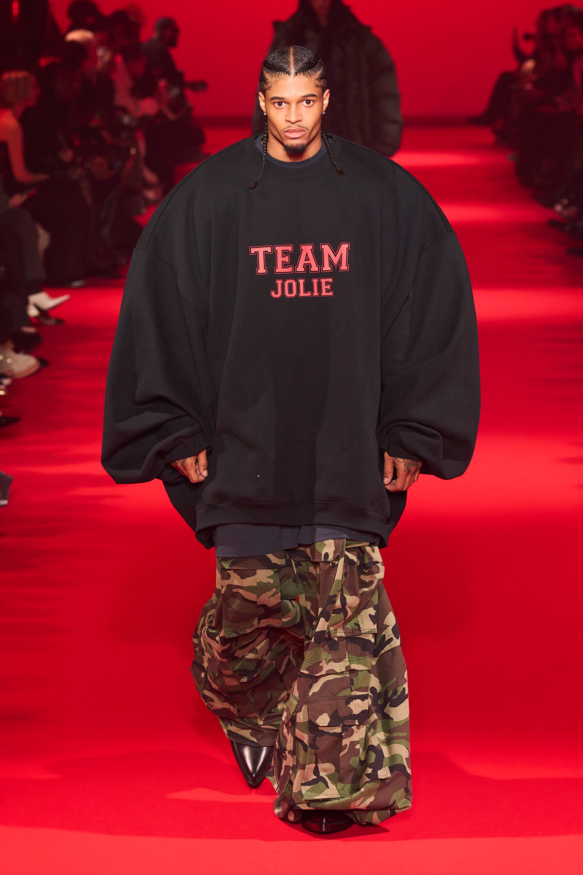Male model walking a red runway, wearing an oversized black sweatshirt with the words 'TEAM JOLIE' and camo cargo pants.