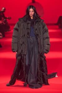 Model walking a fashion runway in a long, oversized black puffer coat against a red-lit background.