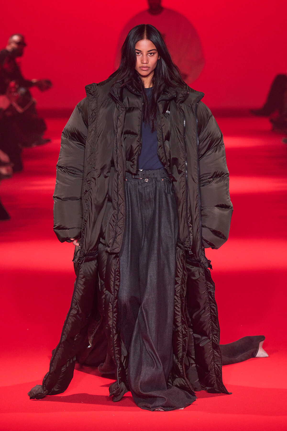 Model walking a fashion runway in a long, oversized black puffer coat against a red-lit background.