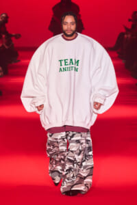Model walking a fashion show runway in an oversized white sweatshirt with green 'TEAM ANISTON' text and camo cargo pants against a red backdrop.
