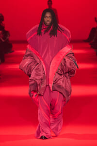 Model walking a fashion runway in a voluminous pink-red padded outfit under red lighting.