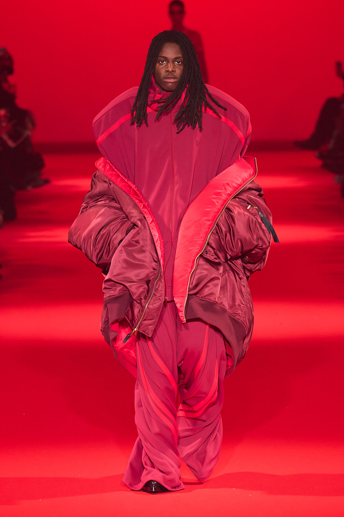 Model walking a fashion runway in a voluminous pink-red padded outfit under red lighting.
