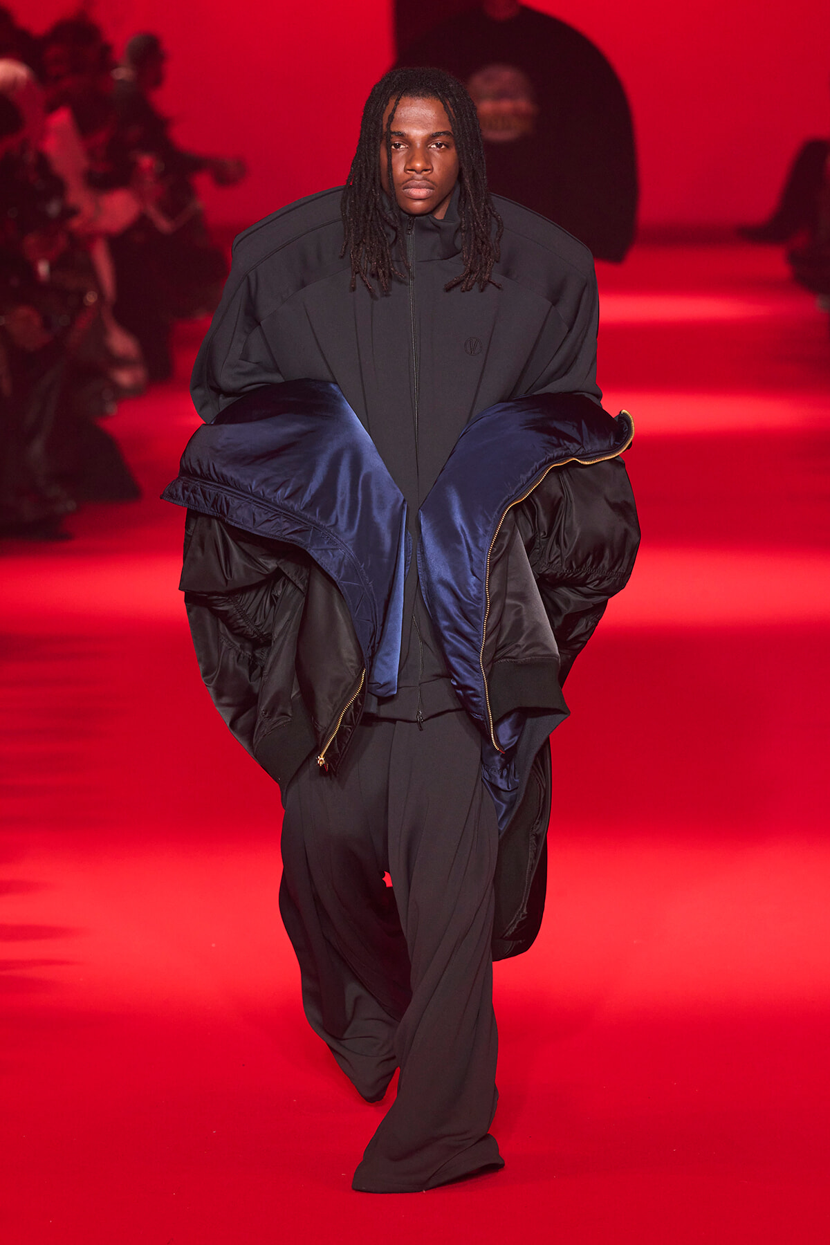 Model walks a red-lit runway in an oversized dark gray coat with a blue puffer draped over the shoulders, creating a bold, layered look,