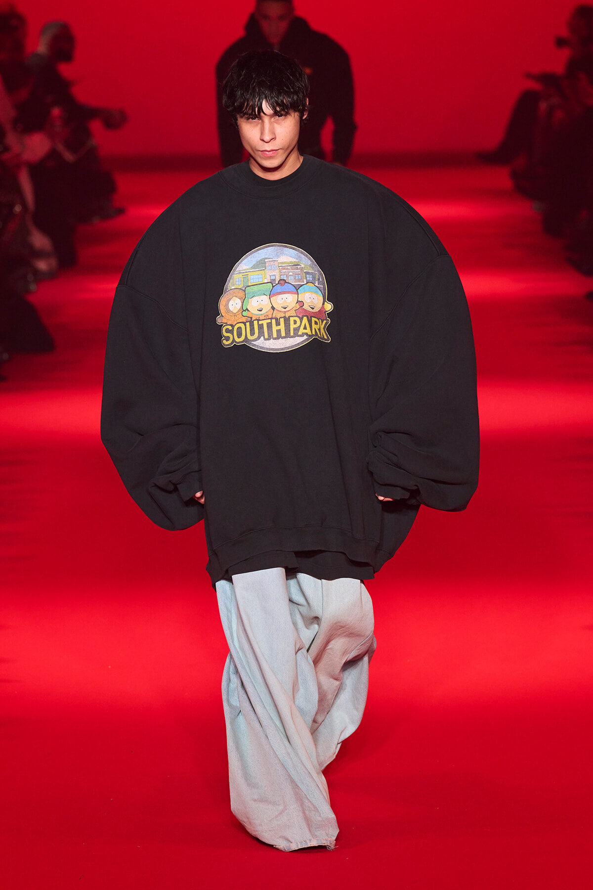 Model on a red-lit runway wearing an oversized black sweatshirt with a South Park graphic.