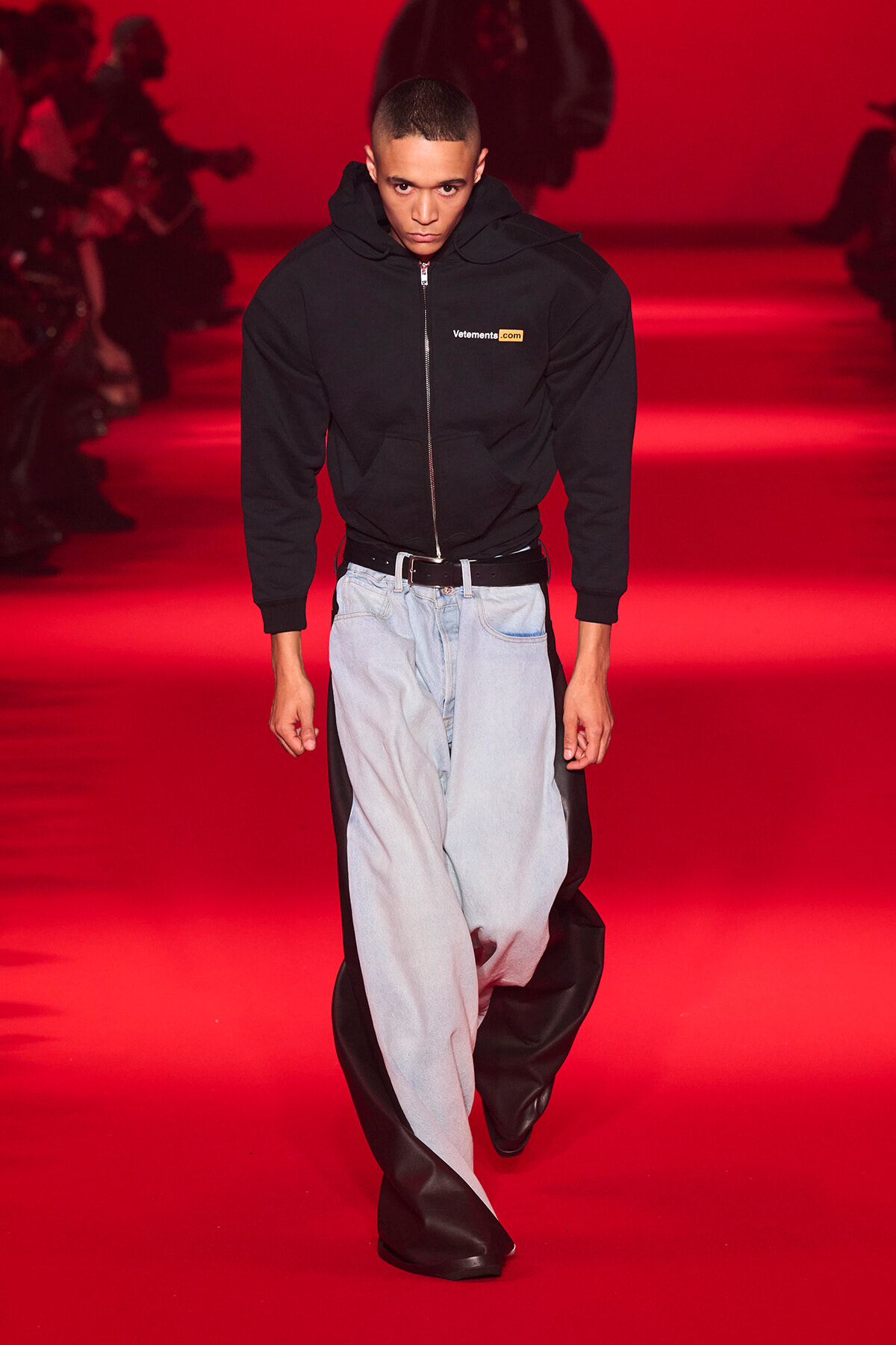 Male model walking a fashion runway in a black zip hoodie with Vetements logo, light-washed jeans, and black leather accents against a red backdrop.