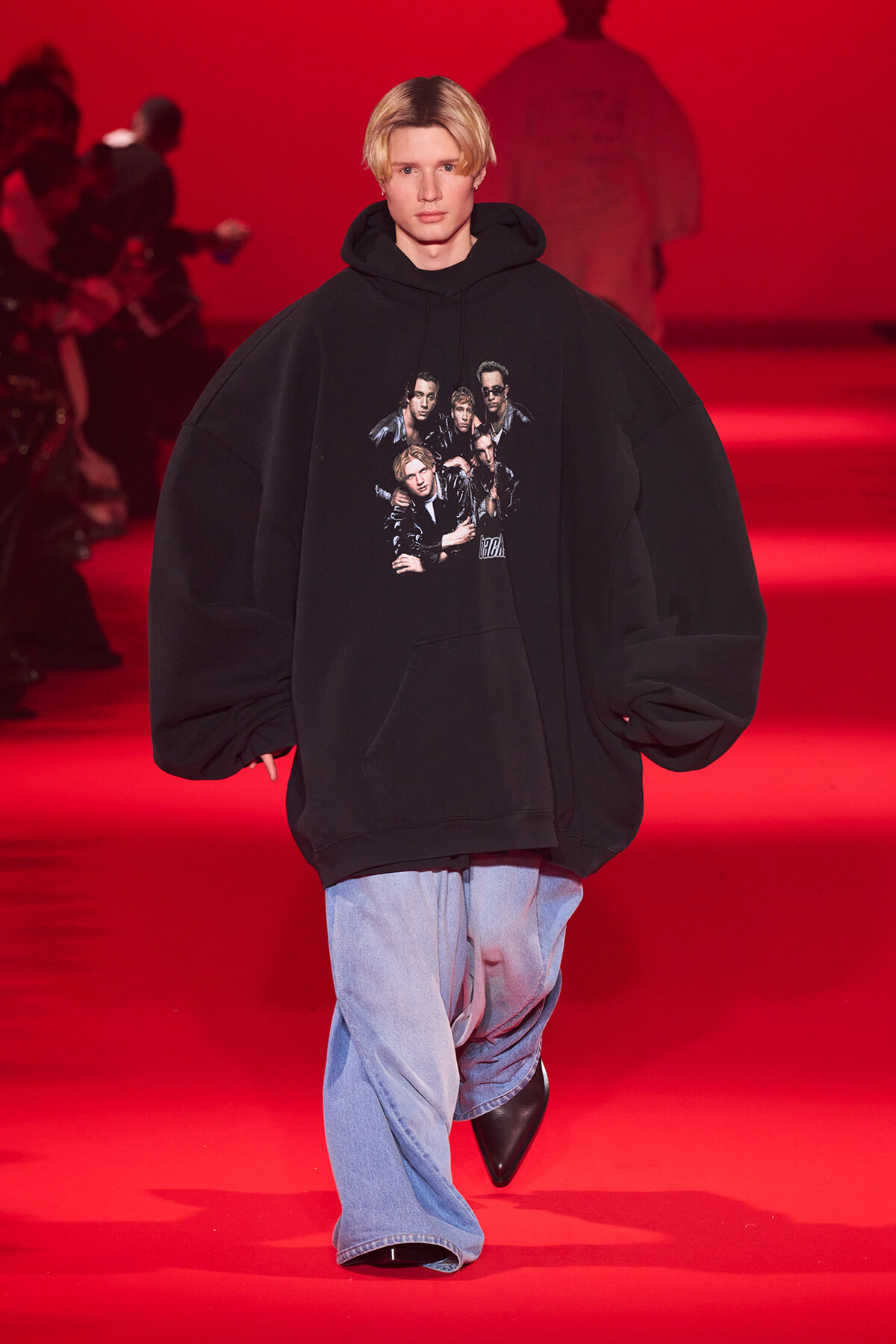 Model walks a red-lit runway wearing an oversized black hoodie with a band/group collage print, paired with light blue jeans.