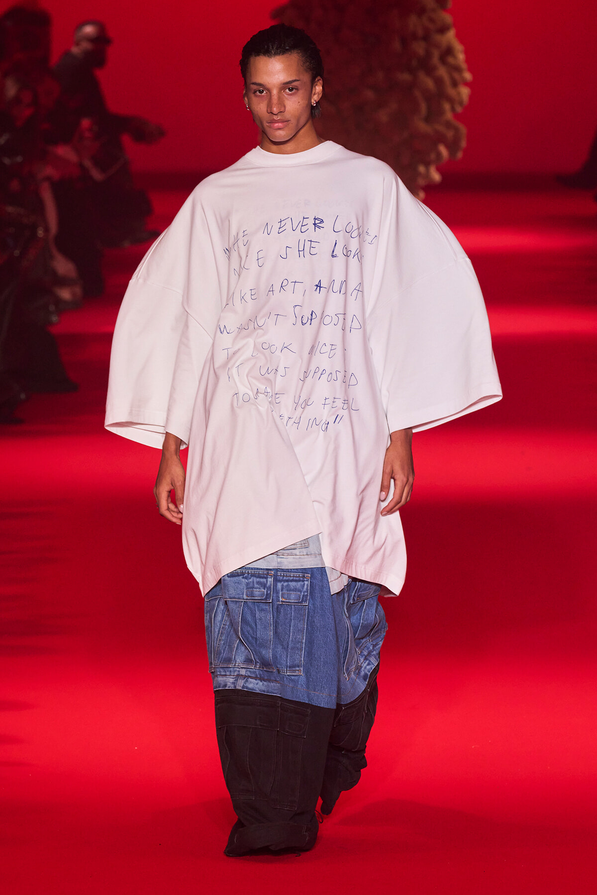 Model on a red-lit runway wearing an oversized white shirt with handwritten text, paired with blue denim cargo pants.