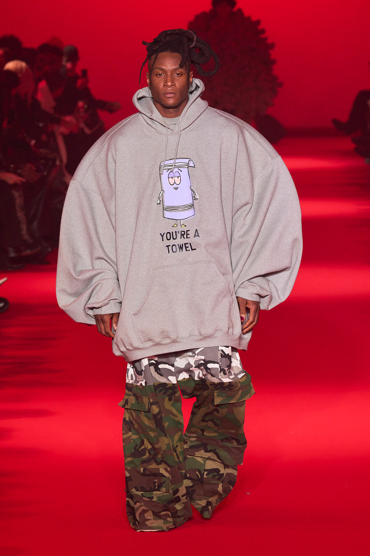 Runway model in an oversized gray hoodie with a cartoon towel graphic and 'YOU'RE A TOWEL' text, paired with camouflage cargo pants under red lighting.