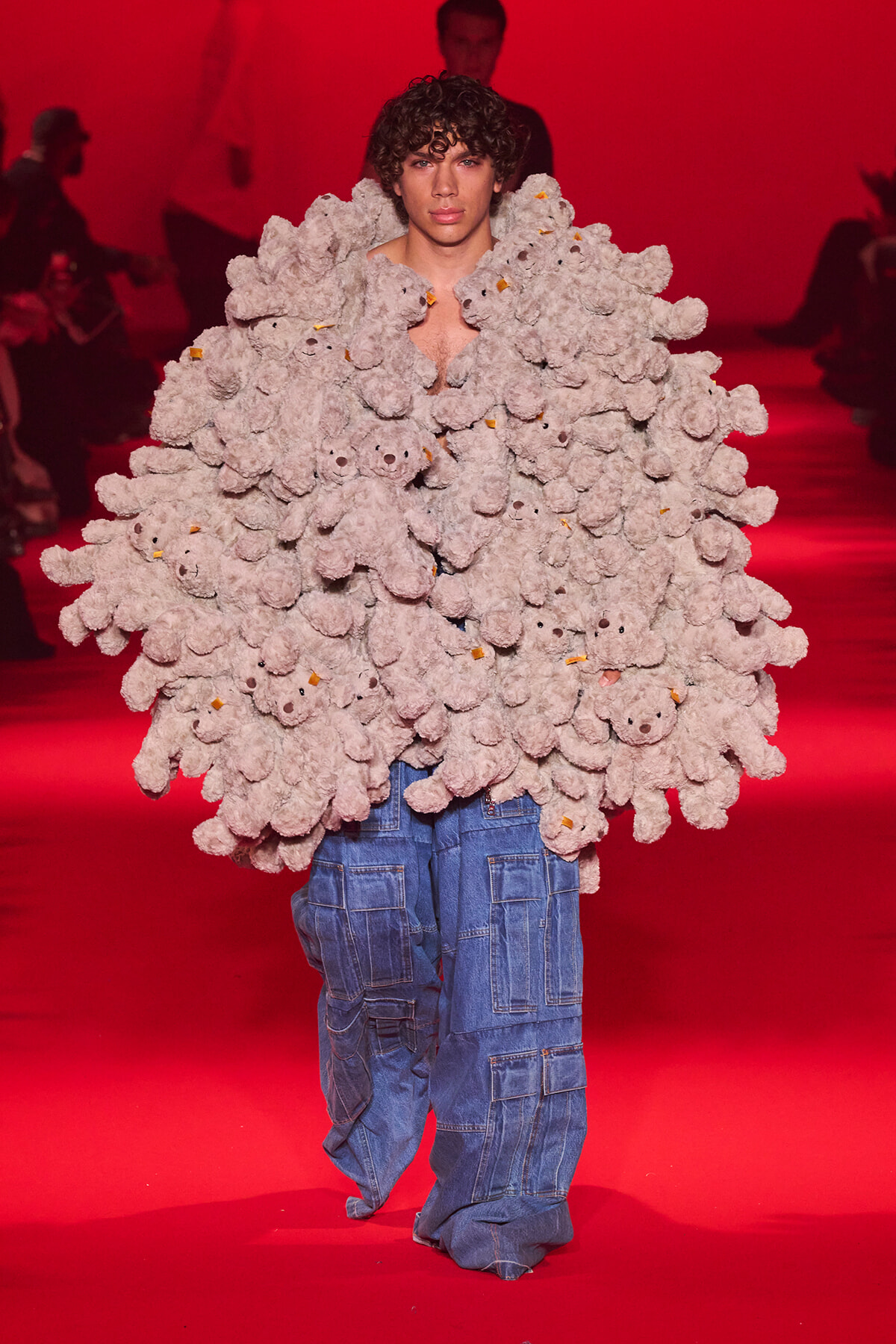 Model on a red runway wearing an oversized gray teddy-bear coat made of many plush bears, paired with baggy blue cargo pants.