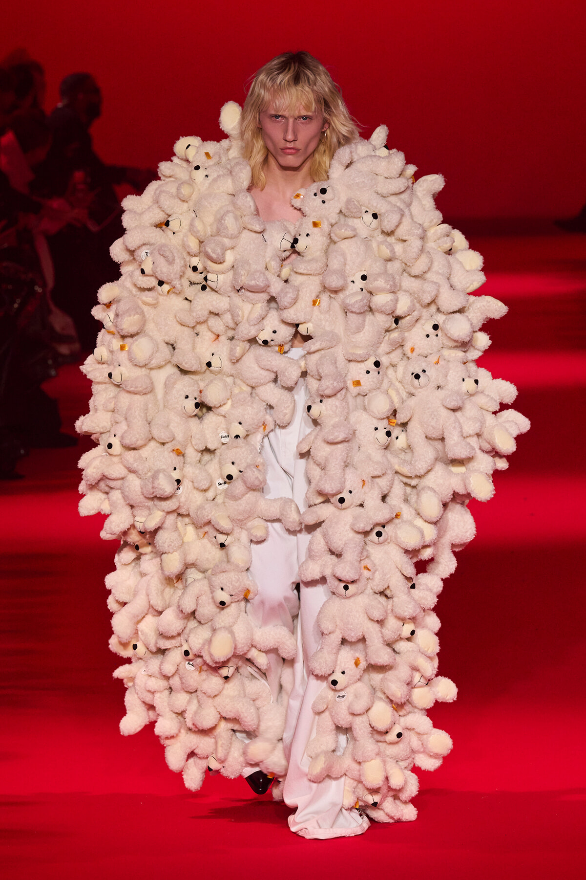 Model on a red runway wearing an oversized coat made of dozens of white teddy bears, creating a fluffy, sculptural look.