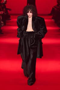Model walking a runway in an oversized black velvet suit with an open chest, under red lighting and a blurred audience in the background.