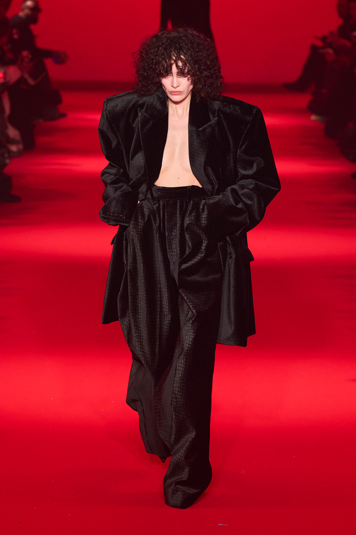 Model walking a runway in an oversized black velvet suit with an open chest, under red lighting and a blurred audience in the background.