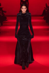 Model walking a long black velvet gown on a red-lit runway during a fashion show.