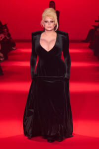 Runway model in a black velvet gown with a deep V neckline walking down a red-lit catwalk, bold makeup, and platinum blonde hair.