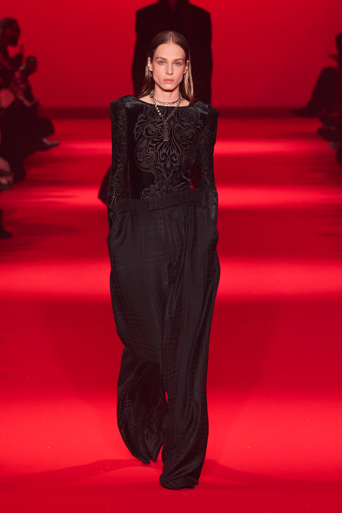 Model walking a runway in a black damask-pattern top and wide-leg pants on a red-lit catwalk, accessorized with a necklace.