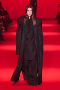Female model walking a red-lit runway in a long black plaid coat, facing forward.