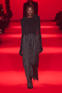 Model walking a red-lit runway in a black glittery top and pinstripe trousers, center stage on the catwalk.