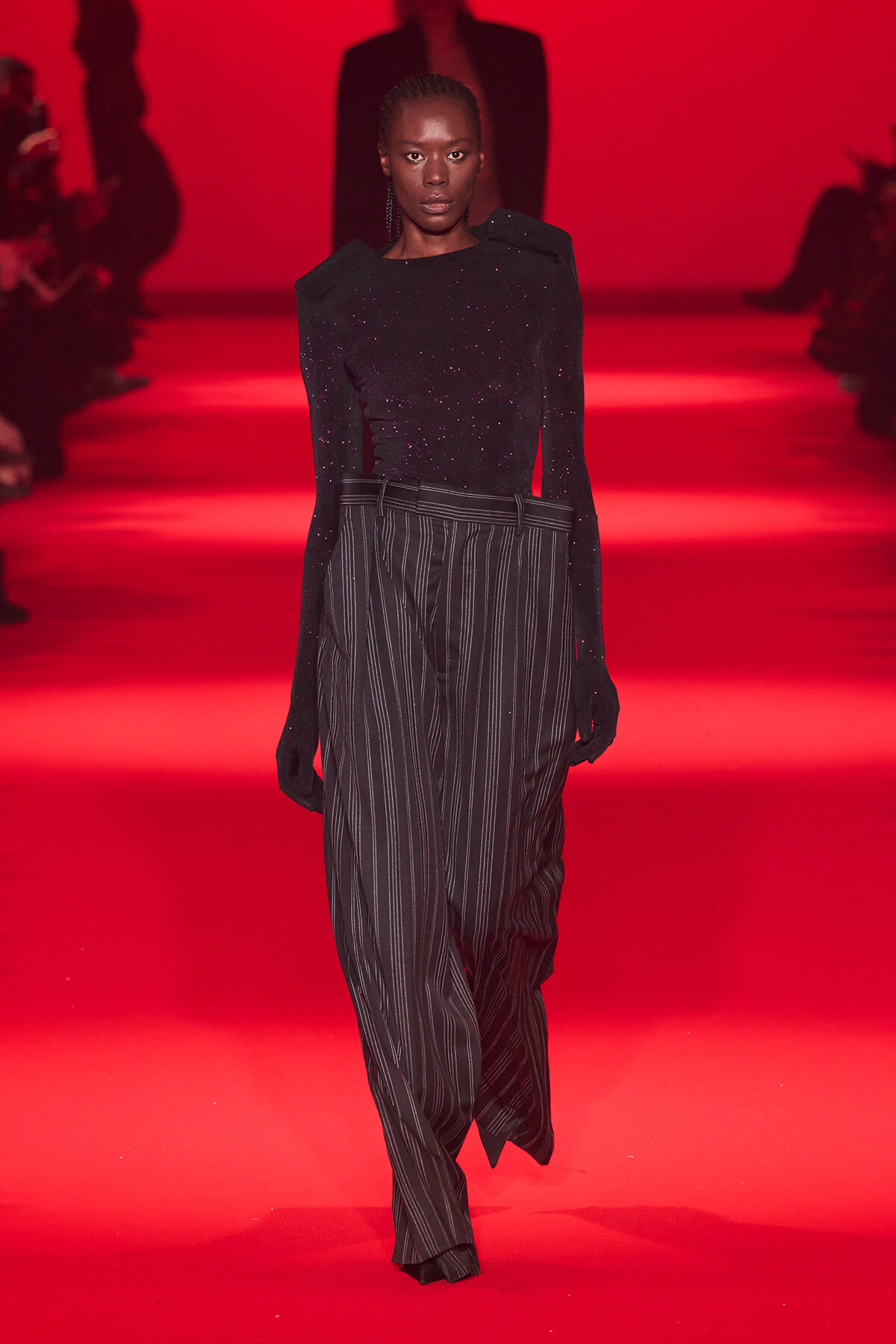 Model walking a red-lit runway in a black glittery top and pinstripe trousers, center stage on the catwalk.