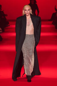 Runway model with blonde hair wearing an oversized pinstripe coat over a nude mesh dress on a red-lit catwalk, looking forward.