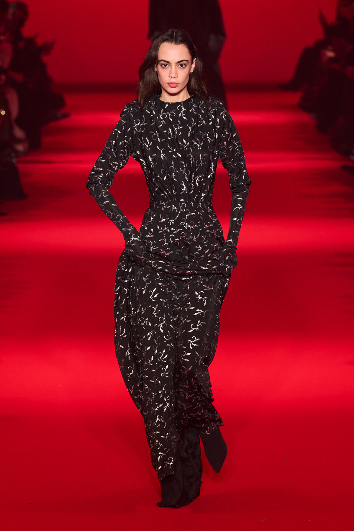Female model walking a red-carpet runway in a black patterned jumpsuit with a gathered waist.