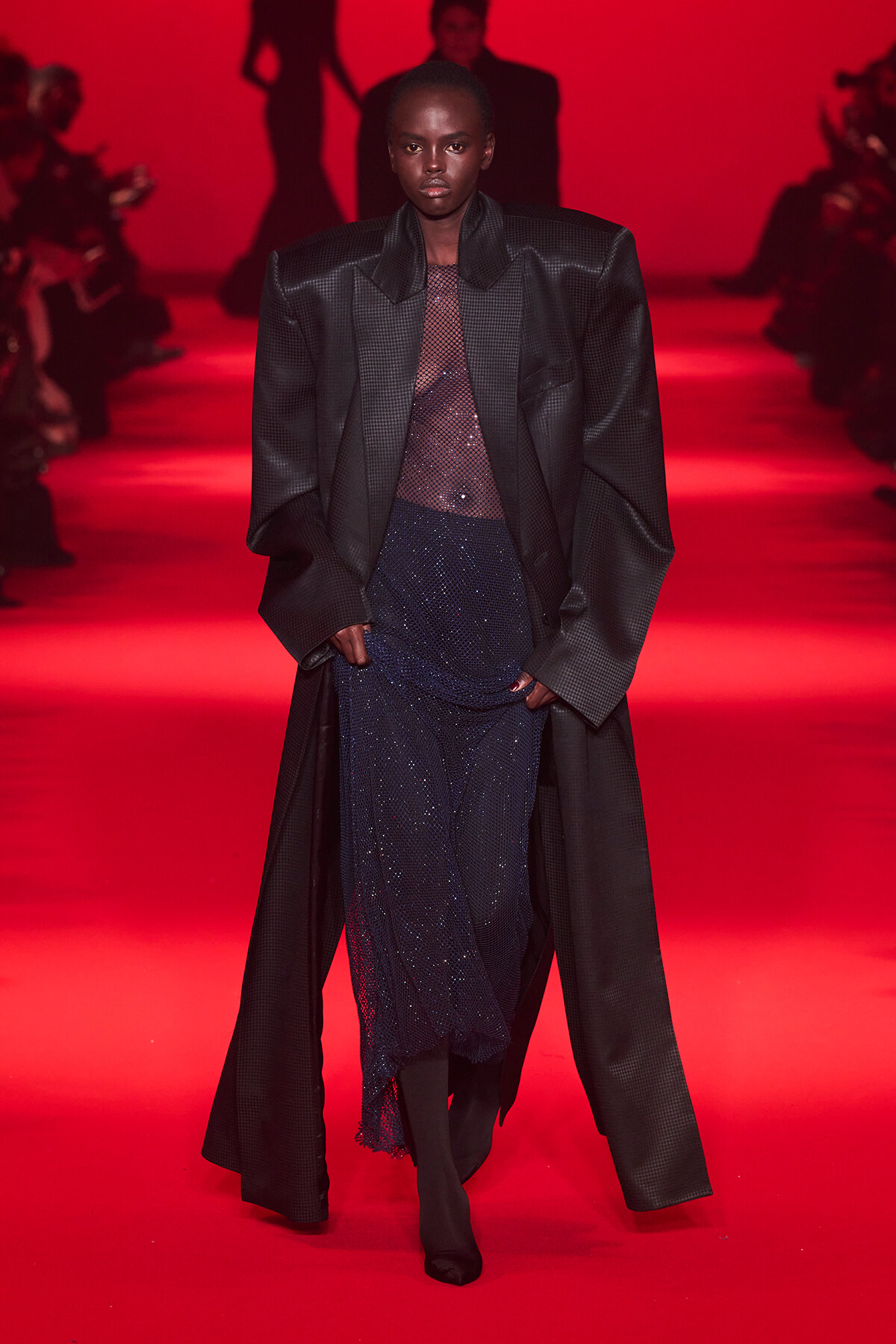 Model walking a runway in an oversized black textured coat over a sheer dotted top and navy sequin skirt, under red lighting, with spectators in silhouette behind.