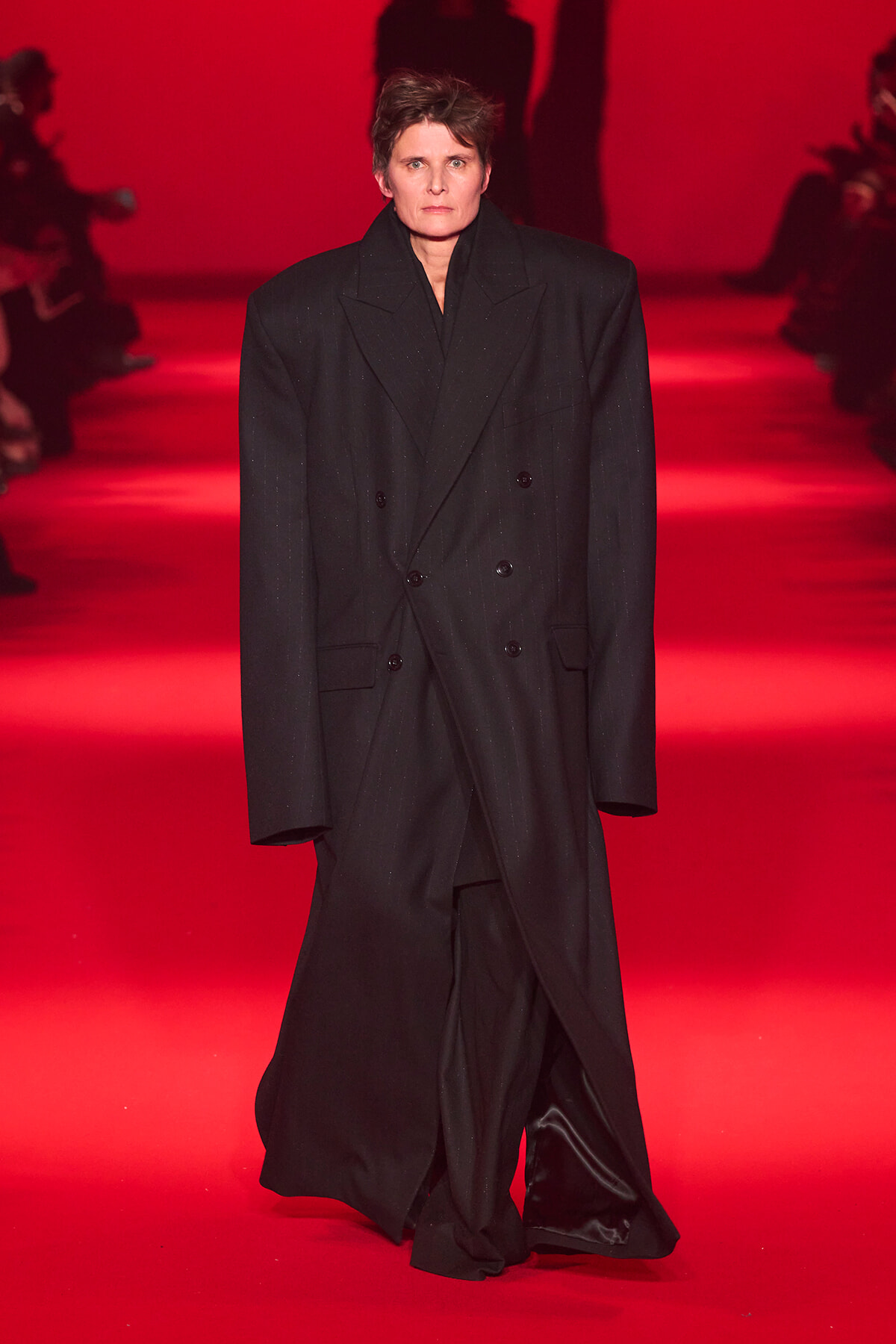 Model walks a runway in an oversized black double-breasted coat on a red-lit catwalk.