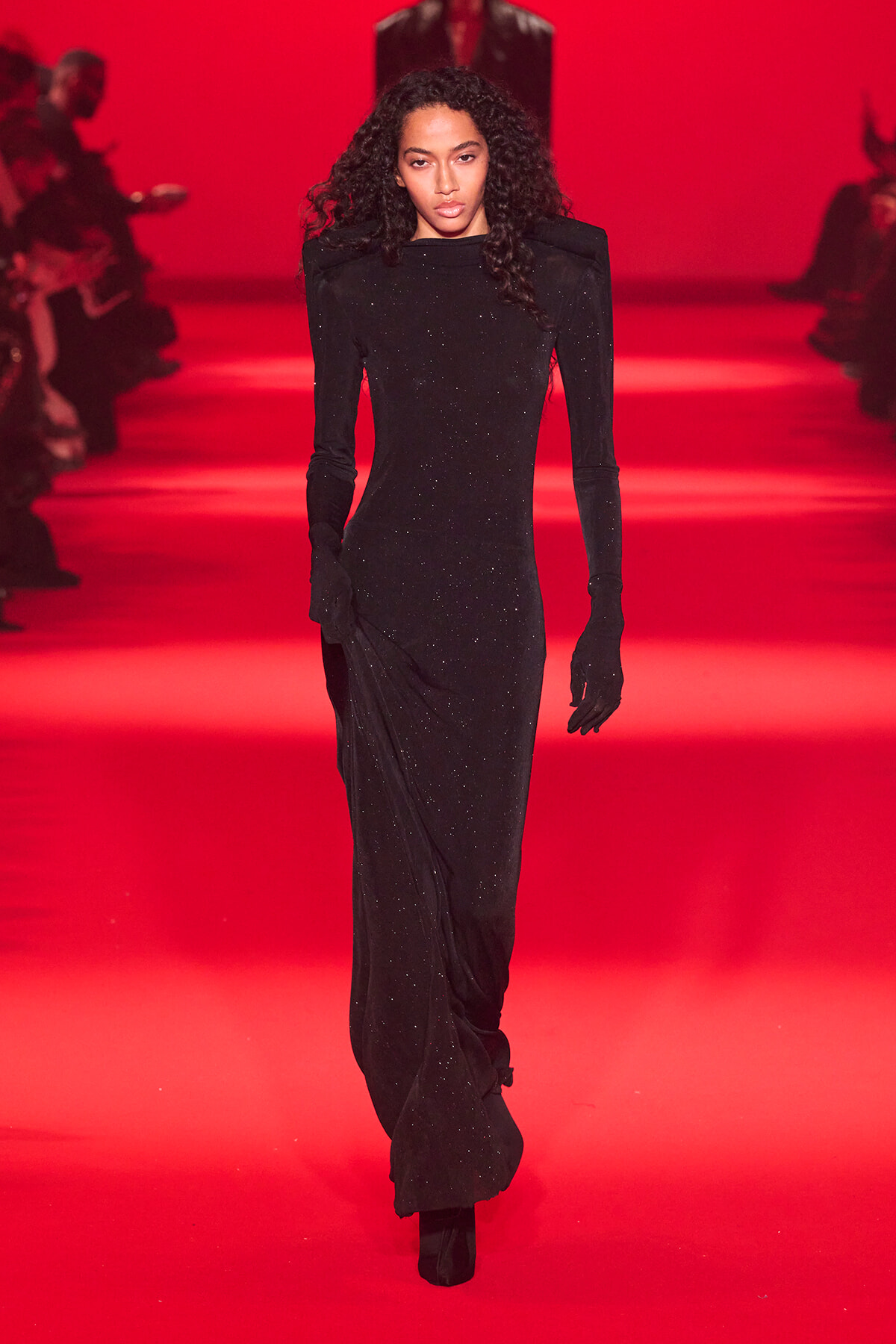 Female model walks a red-lit runway in a long black glittery gown with matching gloves.