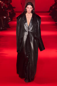 Model walking a red-lit runway in a long, glossy black oversized blazer with a deep V neckline and large shoulders.