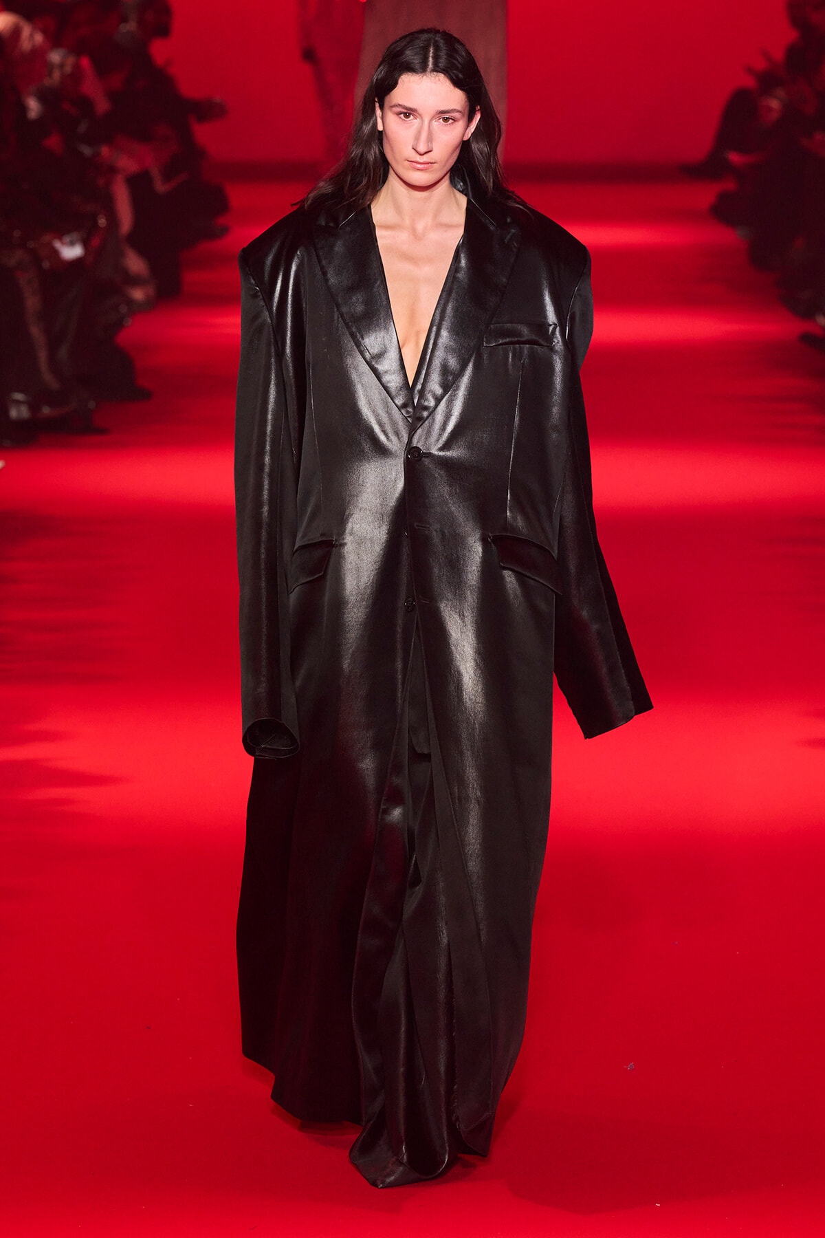 Model walking a red-lit runway in a long, glossy black oversized blazer with a deep V neckline and large shoulders.