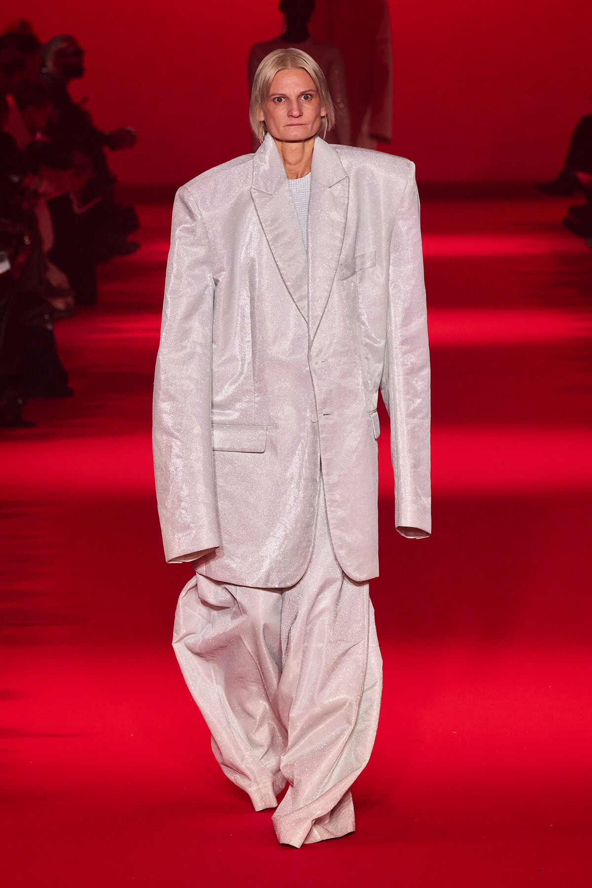 Model walks a fashion runway in a large, shimmering light-silver suit under red lighting, audience silhouettes in the background.