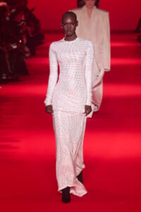 Model walking the red-carpet runway in a long white dotted dress, with another model blurred in the background.
