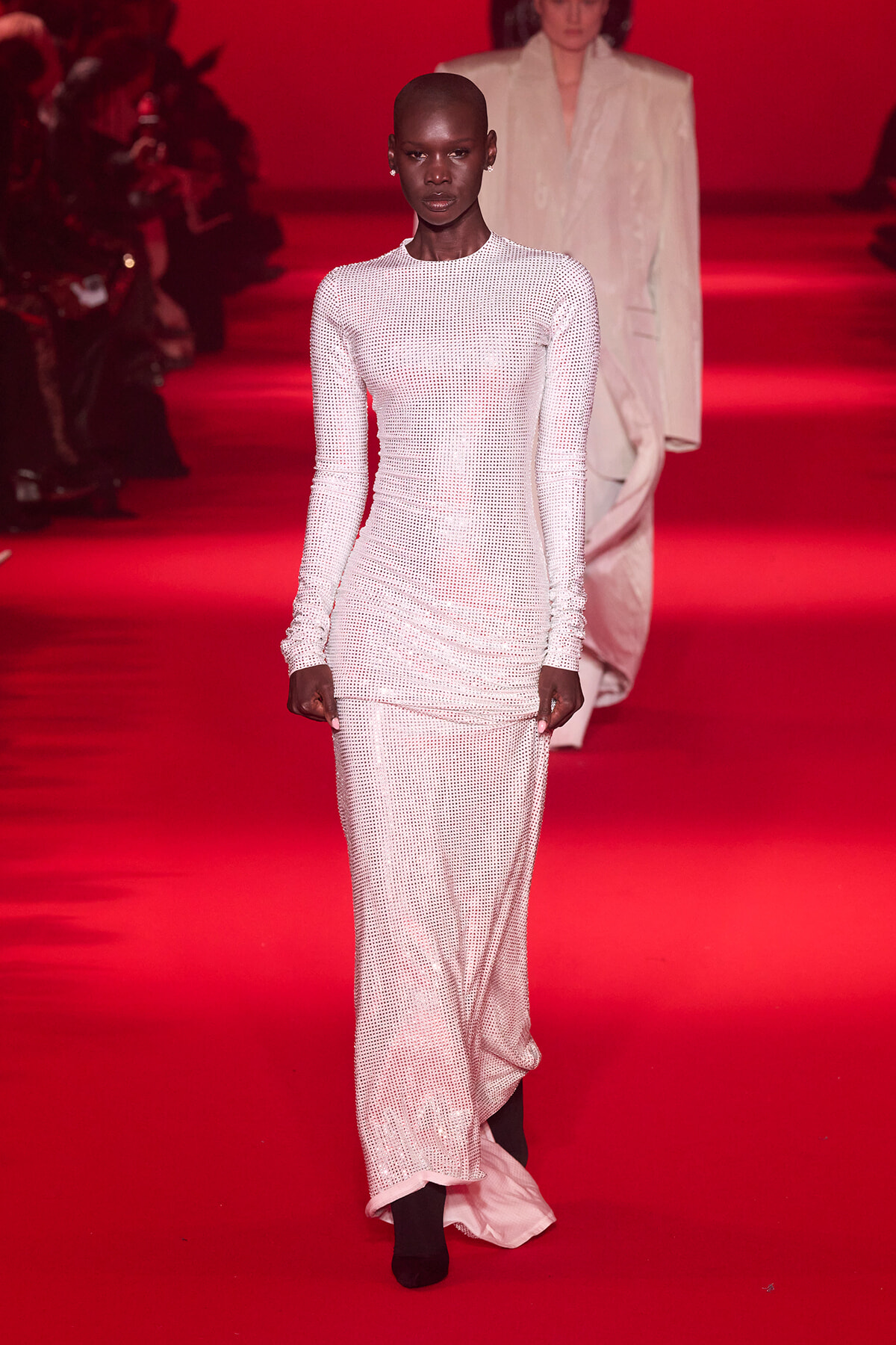 Model walking the red-carpet runway in a long white dotted dress, with another model blurred in the background.