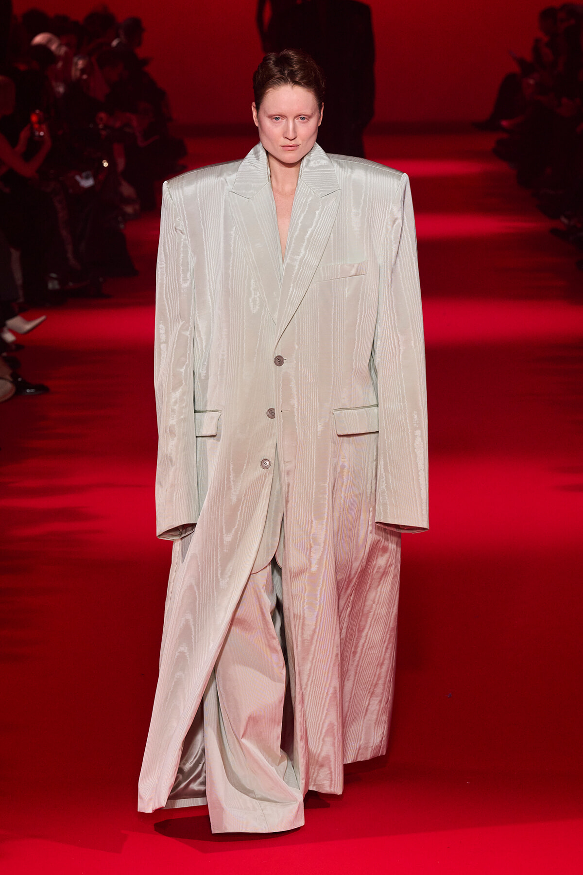 Model walks a red runway in an oversized pale pinstripe suit with broad shoulders and long sleeves, camera facing front.