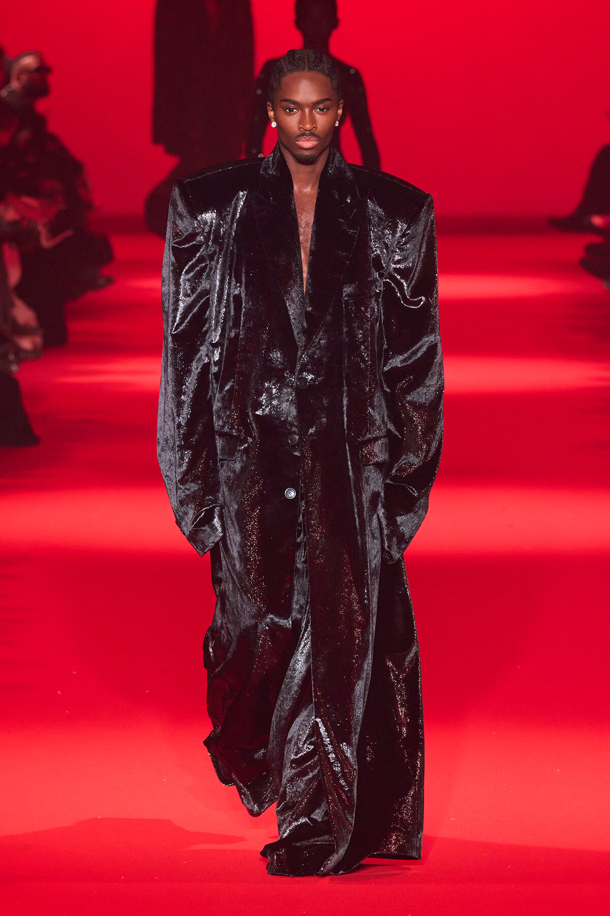 Male model walks a red-lit runway in a long, glossy black oversized coat with broad shoulders.