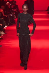 Model on a red-lit runway wearing a full-length black sequined ensemble with long sleeves, posing with hands at her sides.