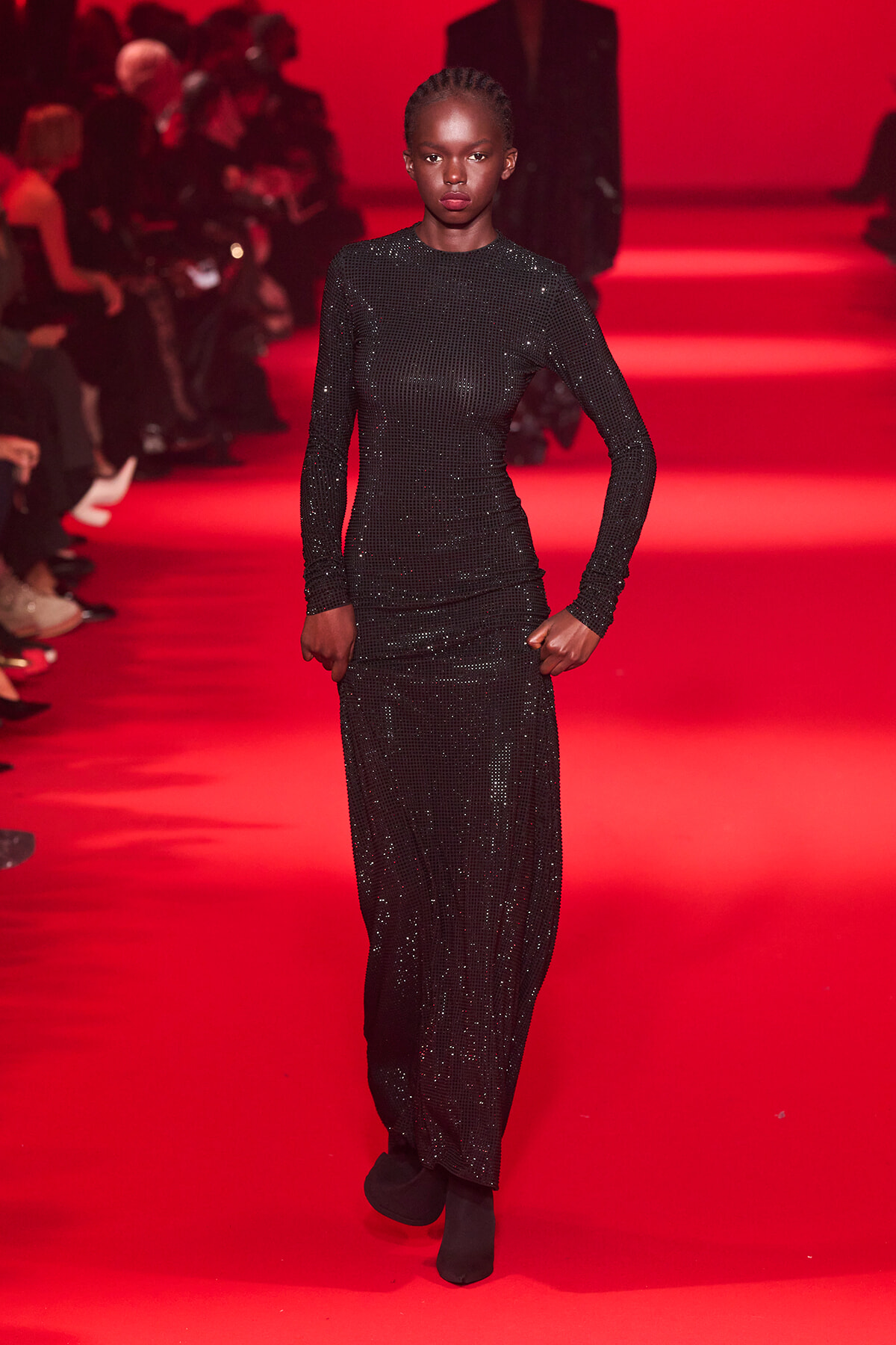 Model on a red-lit runway wearing a full-length black sequined ensemble with long sleeves, posing with hands at her sides.