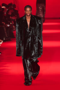 Male model walks the red-lit runway in a glossy black velvet suit with a deep V neckline.