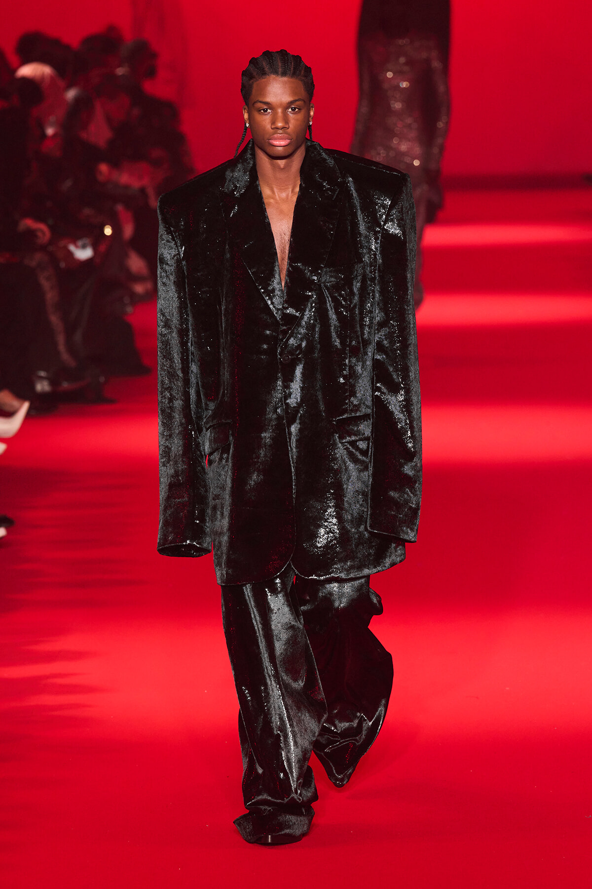Male model walks the red-lit runway in a glossy black velvet suit with a deep V neckline.