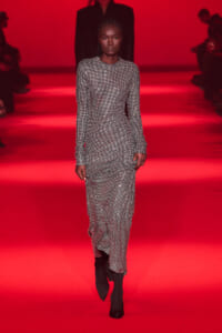 Model walking a fashion show in a long silver-textured gown on a red runway.
