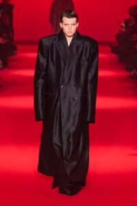 Male model wearing a long, oversized black pinstriped double-breasted suit on a red runway backdrop.
