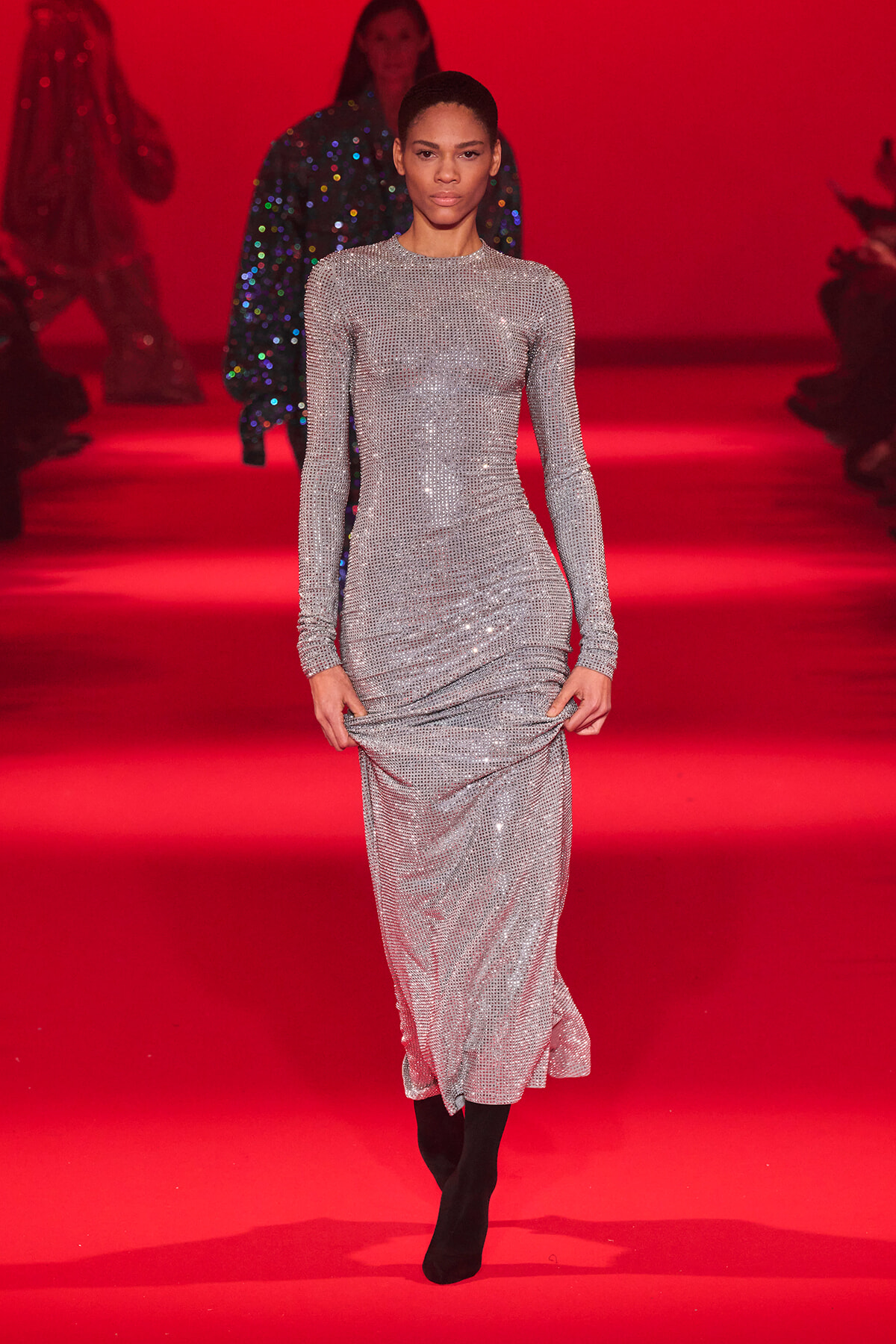 Model walks a silver sequined long-sleeve gown on a red runway show floor, center stage