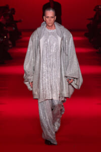 Model walking a runway in a long silver sequined coat and matching pants against a red backdrop.