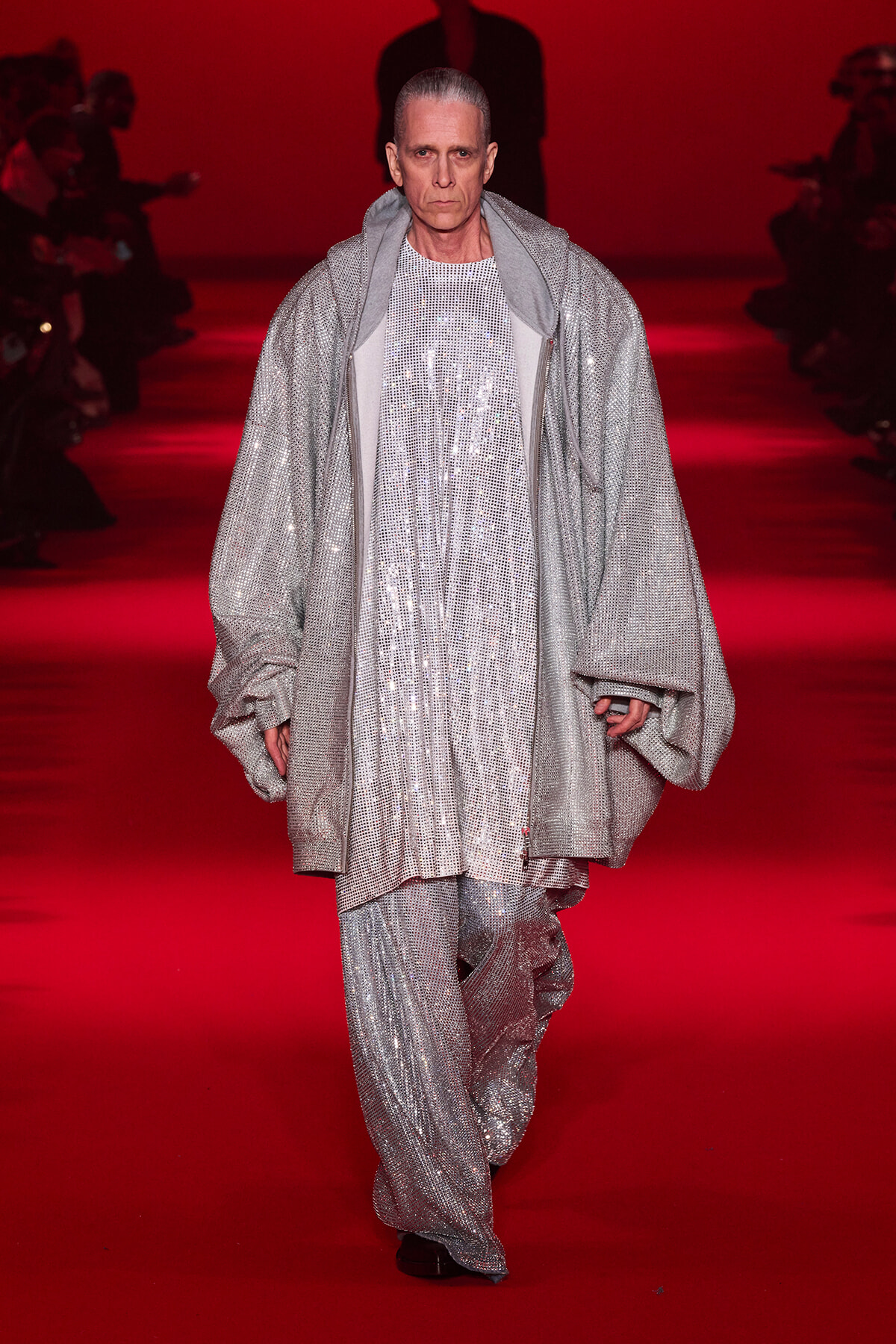 Model walking a runway in a long silver sequined coat and matching pants against a red backdrop.