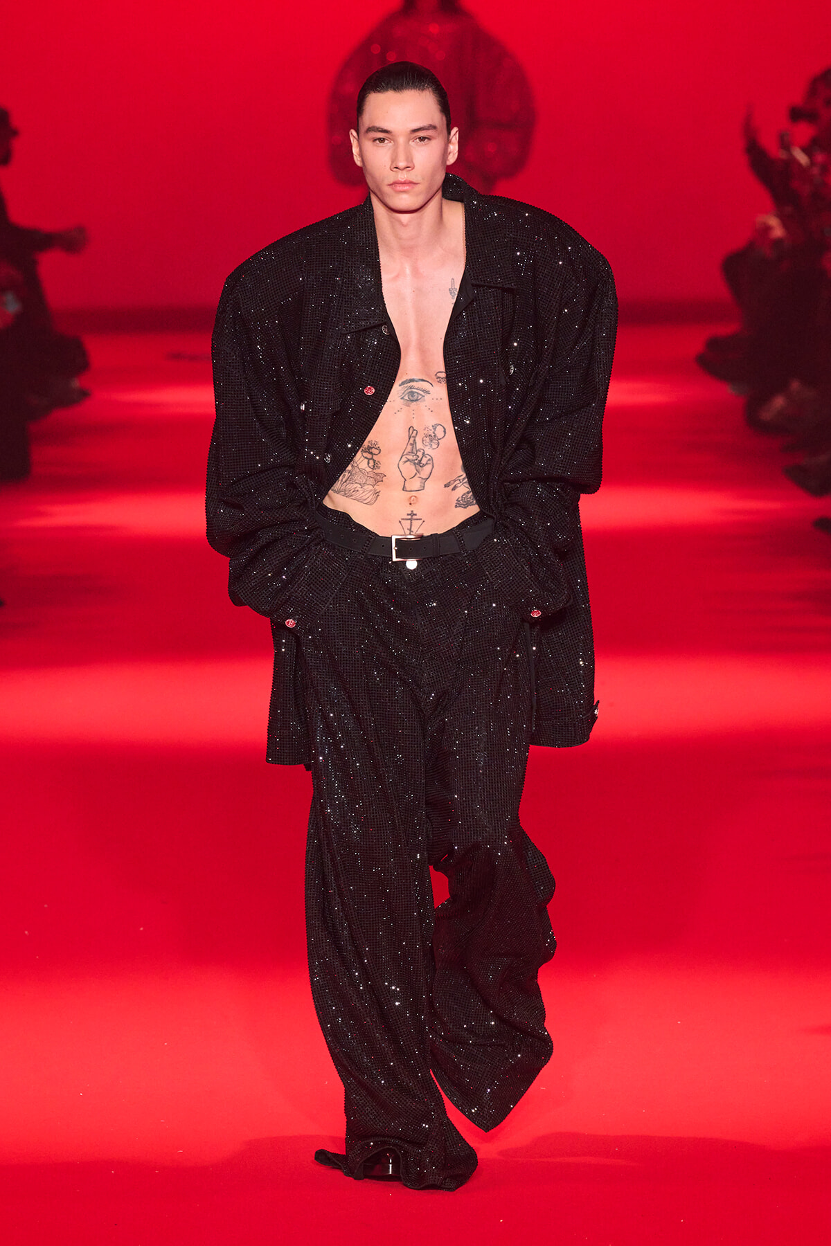 Male model walking a red-lit runway in a black sequined suit with an open chest showing tattoos.