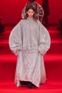 Model on a red runway wearing a glittering silver cape-like dress with oversized sleeves.