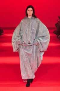 Model walking a fashion show in a shimmering silver poncho-style dress with oversized sleeves on a bright red catwalk, hands in front holding fabric.